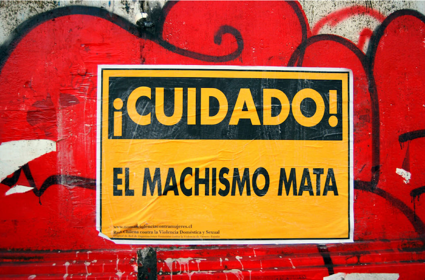 Alt Text: Poster on a street wall that reads: "Caution! Machismo kills". Behind the poster, red graffiti can be seen, though illegible.