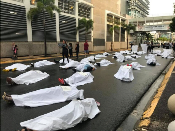 Alt Text: Multiple women (at least 13) are lying down in the street with white sheets covering them. On top of those sheets are pieces of paper/cardboard with names of gendered violence written on them.
