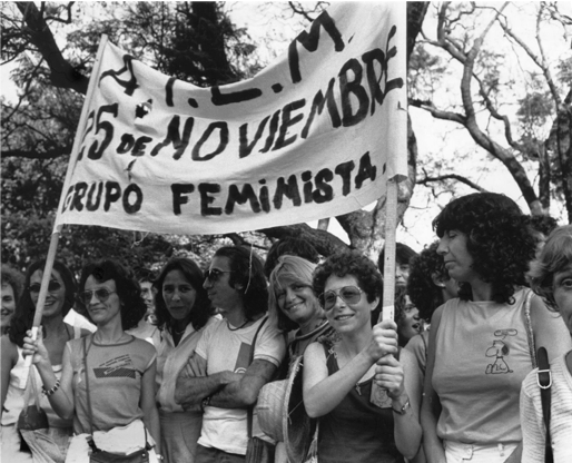Alt Text: Various smiling people (which are mostly women) marching in what appears to be a protest for women's rights. Two women are holding sign saying: 25 of November, Feminist Group.
