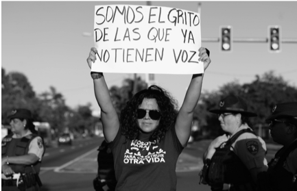 Alt Text: Woman in the street in front of a line of cops. She is holding a sign that reads: "We are the screams of those that no longer have a voice"
