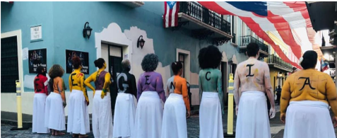 Alt Text: Row of women standing on a street that leads to the "Fortaleza" (similar to the White House in the US). Each woman is painted in a specific color with a letter painted on their backs. Together they spell "emergency". It represents the state of emergency the feminist groups are asking for in response to the spike in gendered violence on the Island.