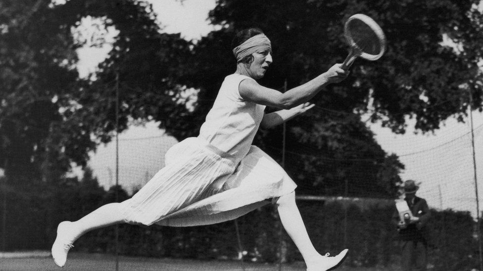 Suzanne Lengen in 1920’s tennis outfit. All white outfit, skirt to her knees, and shirt that covers her shoulders and white shoes.
